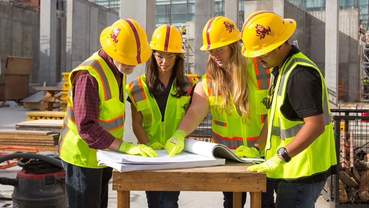 A team of three students and their instructor are gathered around a large pad of paper while at a construction site wearing safety gear.